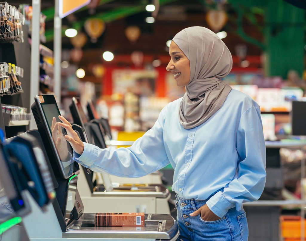 A smiling Muslim woman checks her item out at a self-checkout lane in a store.